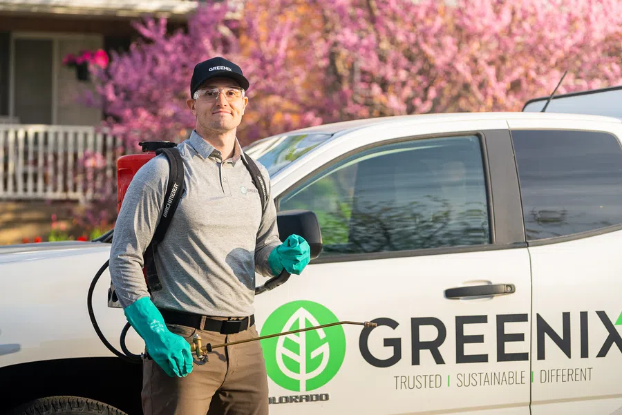 Pest control technician in gloves stands beside Greenix branded truck with blooming pink trees in background