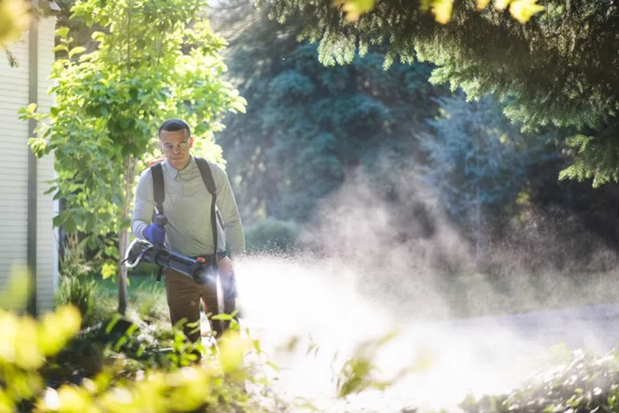 Man using a backpack leaf blower to clear leaves and debris in a sunny garden yard.