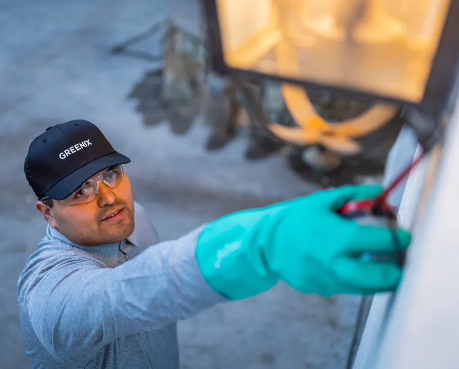 Technician wearing safety glasses and green gloves inspecting outdoor lighting fixture at dusk.