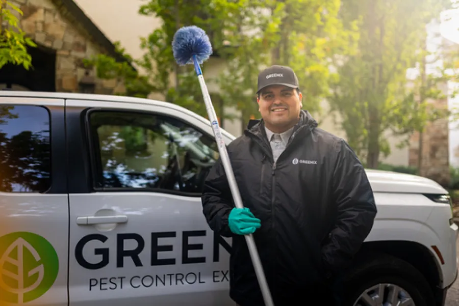 Pest control technician holding a blue brush standing in front of a Greenex company truck outdoors.