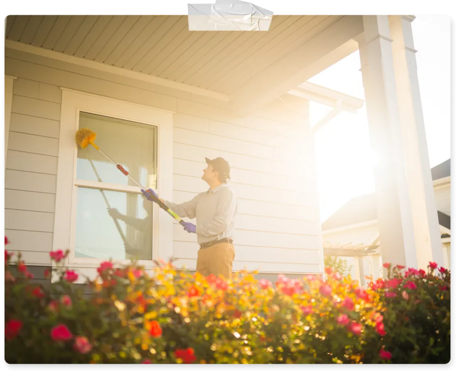 Man cleaning a house window with a long-handled duster in bright sunlight and colorful garden flowers.