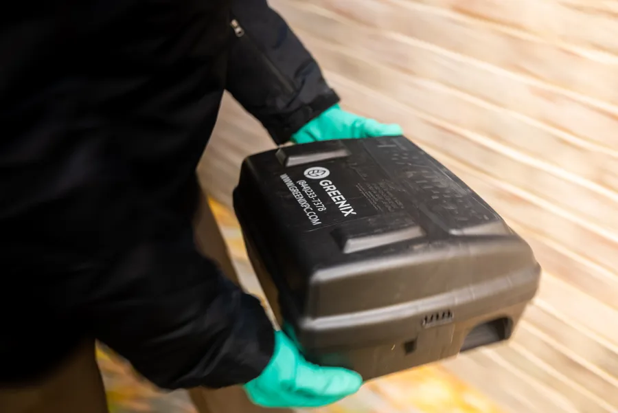 Person wearing green gloves holding a black Greenix tool case against a blurred background.