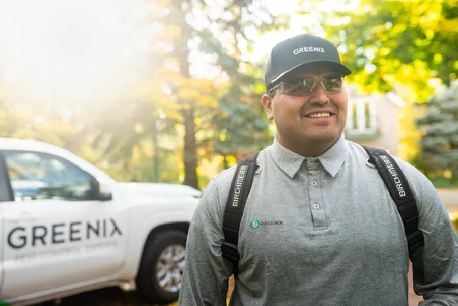 Smiling pest control worker wearing Greenix cap and uniform stands outdoors near a branded company truck in a sunny neighborhood.