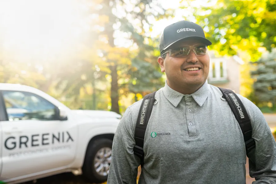 Smiling pest control technician in Gray shirt and cap with Greenix logo standing beside company truck outdoors