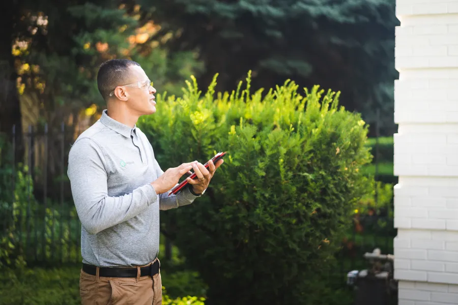 Male technician using tablet outdoors near green bush and white brick wall in bright daylight.