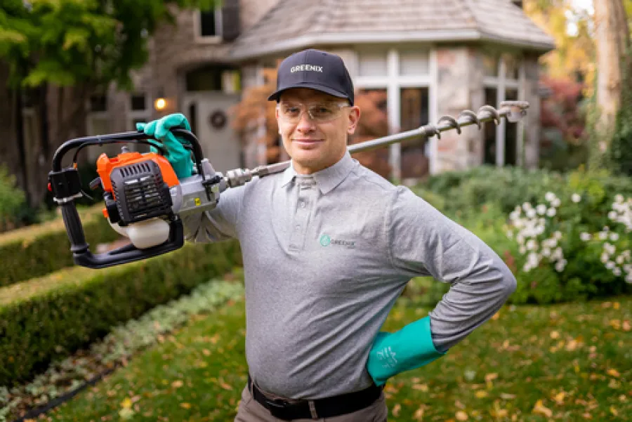 Man in gloves and cap holding a gas-powered garden auger in a residential garden with greenery and a house in the background