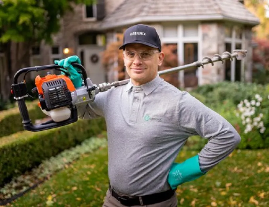 Man in grey shirt and gloves holding an earth auger in a garden with hedges and flowers