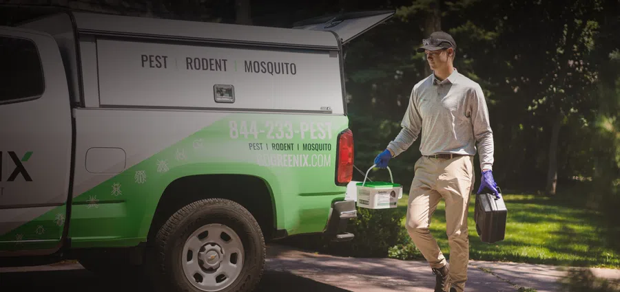 Pest control technician carrying equipment from a green service truck in a residential area.