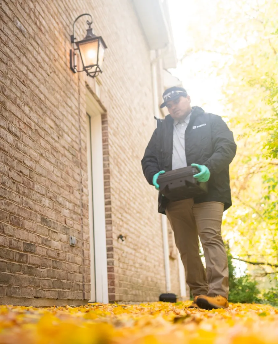 Person wearing gloves and jacket holding a black device walking on yellow autumn leaves beside a brick house.
