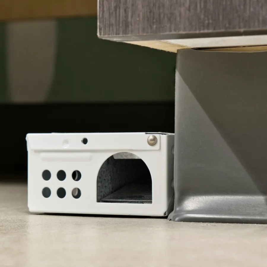 White metal rat trap placed under a wooden cabinet on a tiled floor with holes on its side.