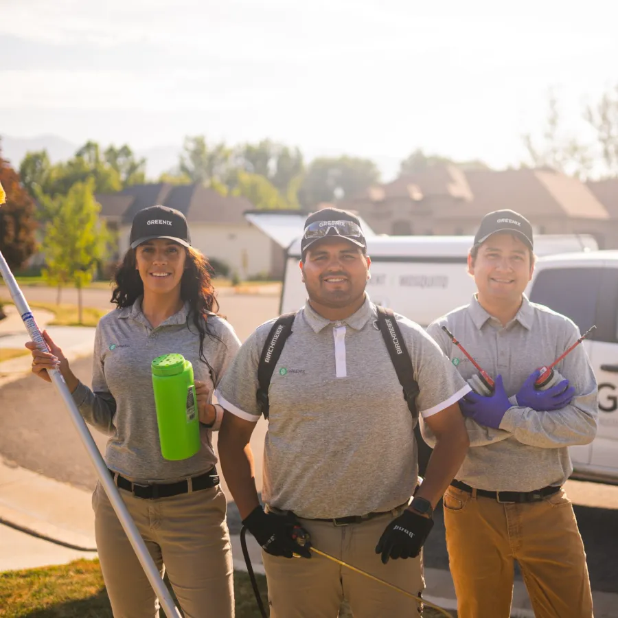 Three pest control technicians in uniform standing outdoors with equipment and company vehicle in background.