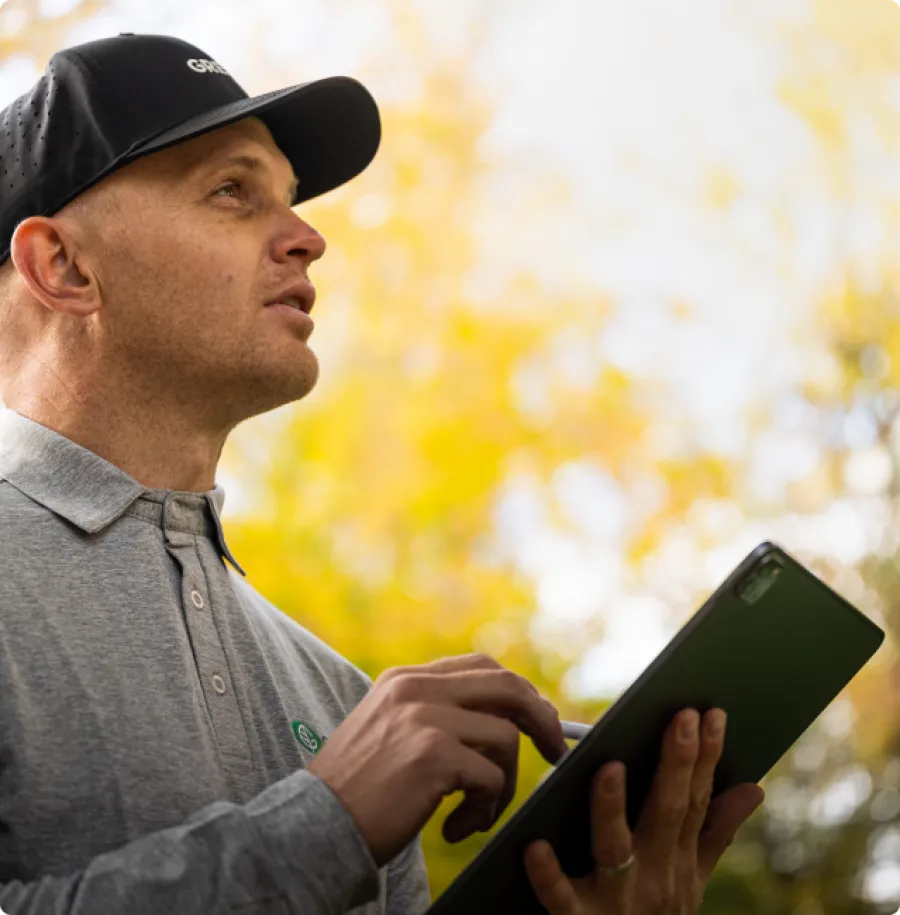 Man in gray shirt and black cap using tablet outdoors with blurred yellow foliage background.