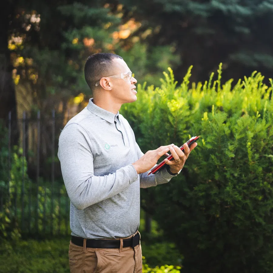 Man in safety glasses and grey polo using a tablet outside near green bushes and trees during daytime