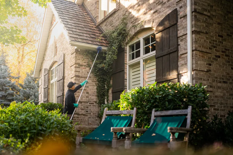 Person cleaning house exterior brick walls with a long brush during daylight surrounded by green plants and chairs.
