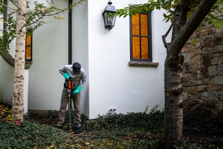Person using a power auger to drill into ground near white house wall with green ivy and two trees.