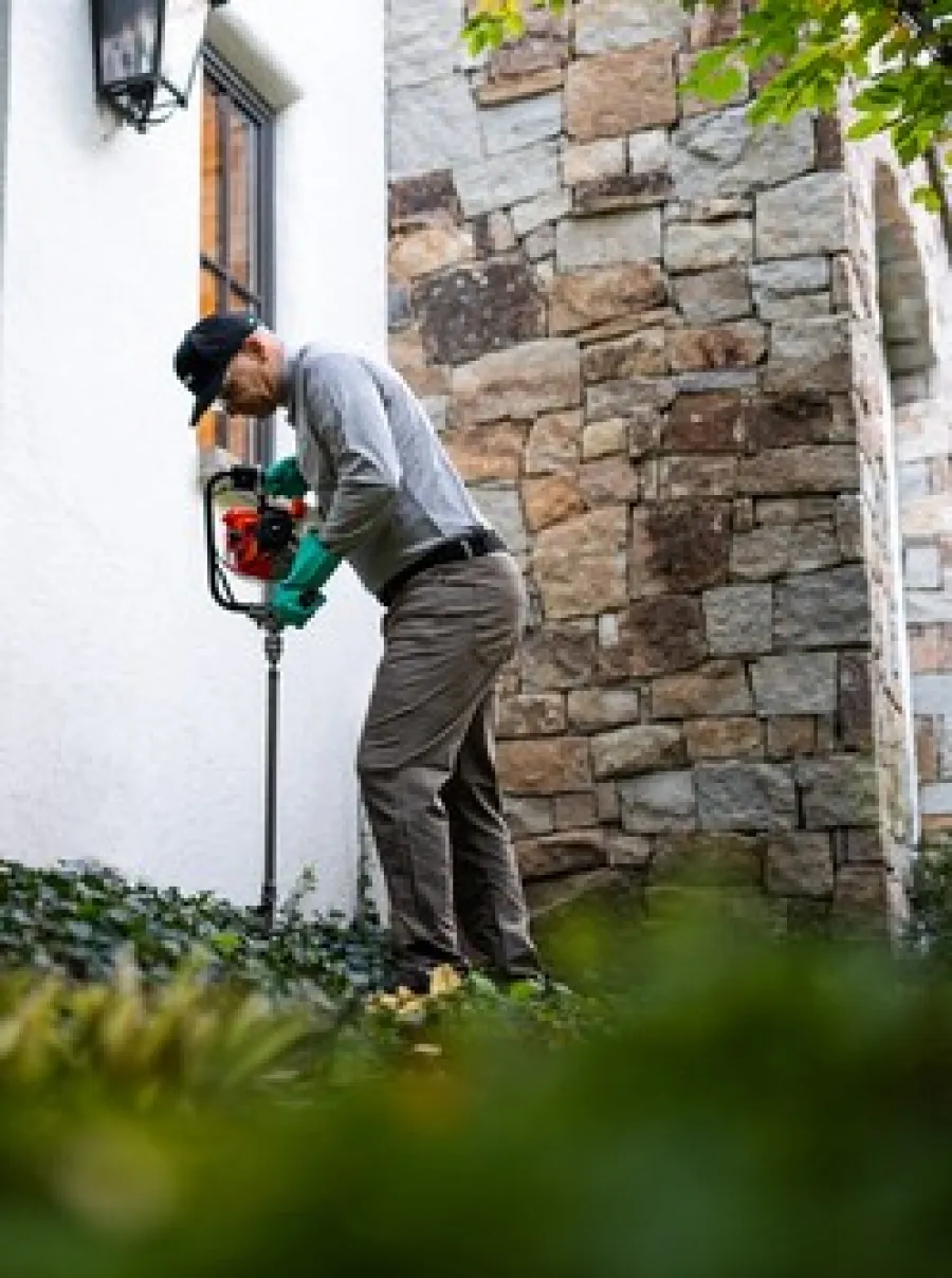 A man using a garden auger in a landscaped yard near a stone wall.