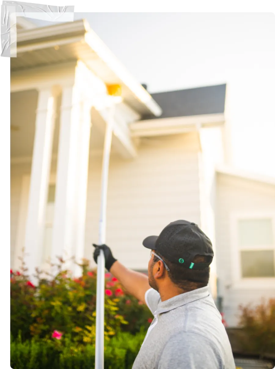 Man in cap using a long paint roller to paint the exterior white wall of a house in daylight.