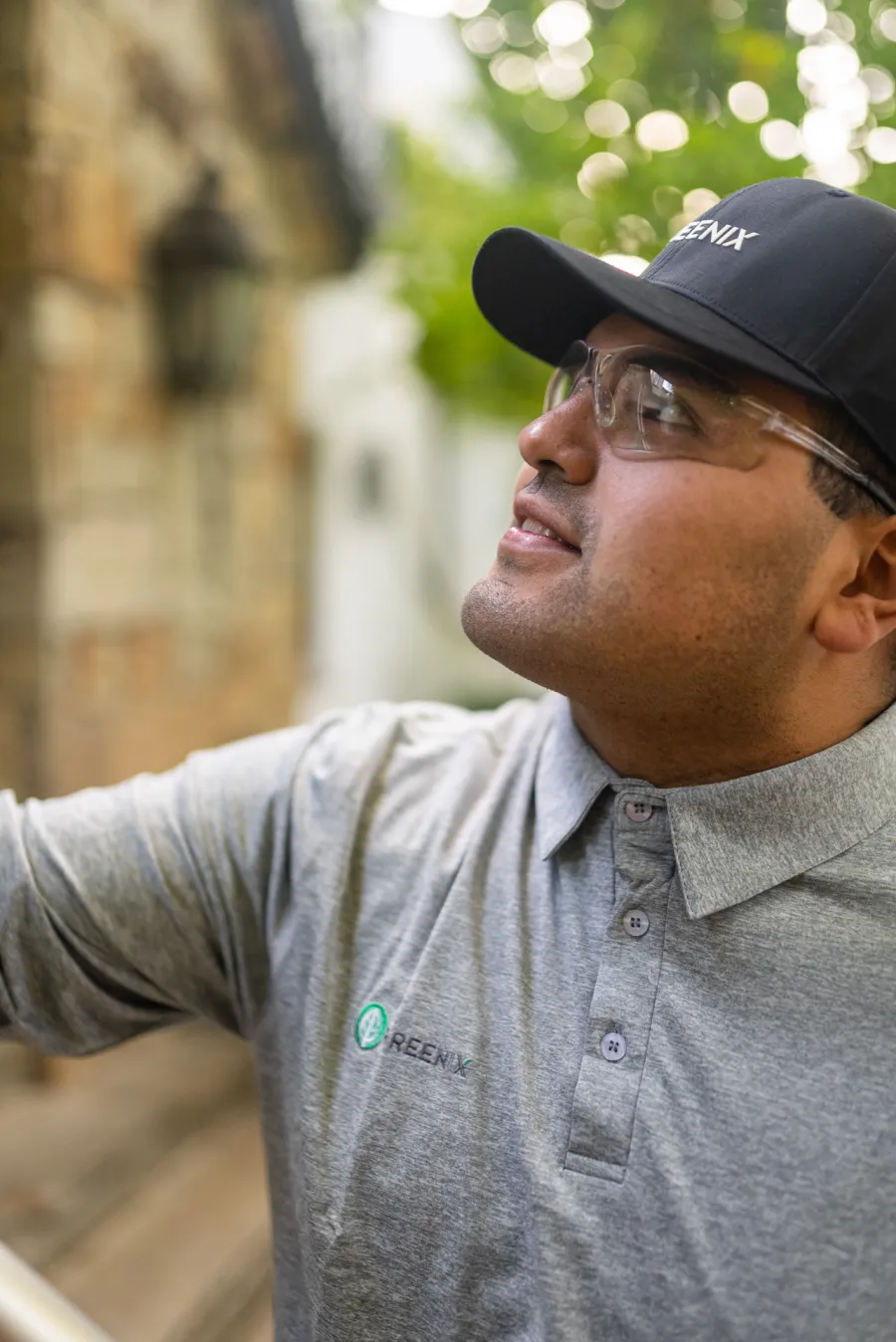 Man wearing safety glasses and a black cap inspecting outdoors, dressed in a gray polo shirt with a logo.