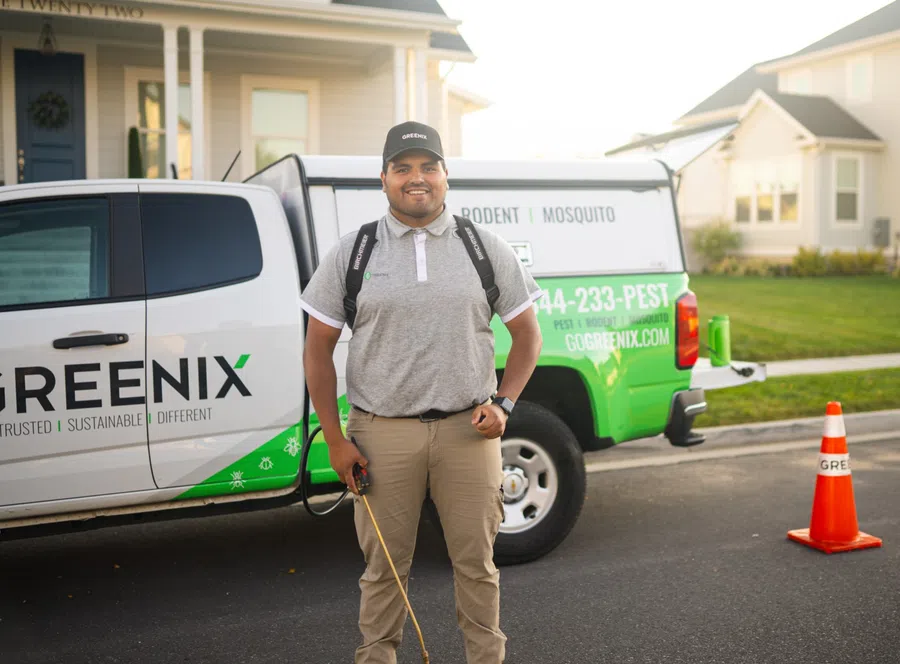 Smiling pest control technician in uniform standing by Greenix branded service truck in a residential neighborhood.