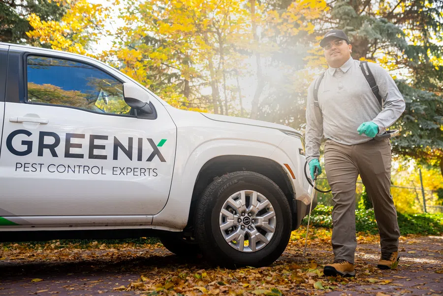 Pest control technician walking with equipment near a Greenix Pest Control Experts vehicle in autumn setting.
