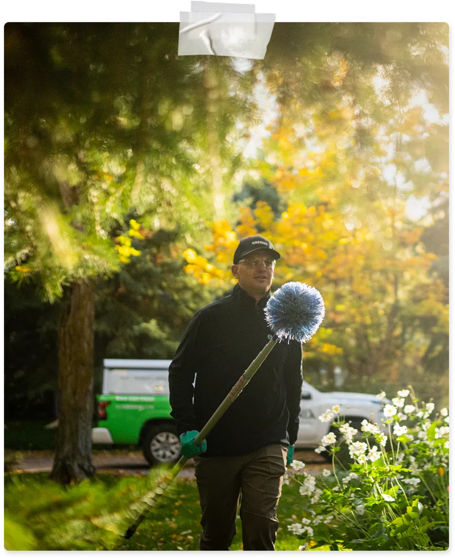 Man wearing a cap holding a long-handled brush in a garden with green and yellow foliage and a vehicle in the background