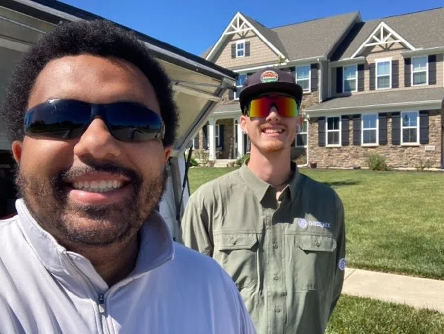 Two men wearing sunglasses smiling outside a large suburban house on a sunny day.
