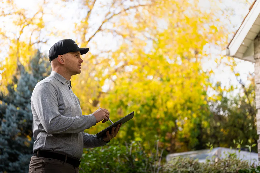Man in gray shirt and black cap using tablet outdoors with autumn trees and house in background