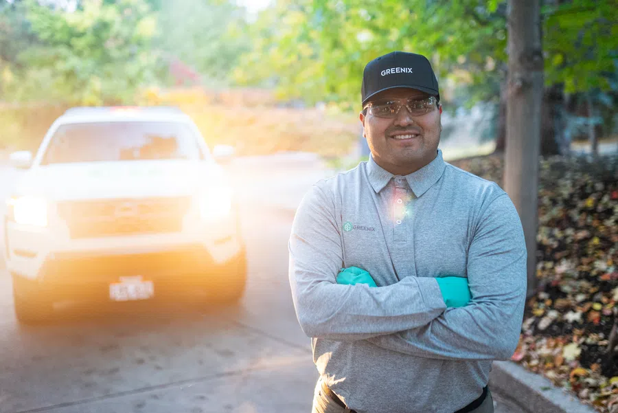 Smiling man in Greenix uniform and cap stands with arms crossed near white vehicle on tree-lined driveway.