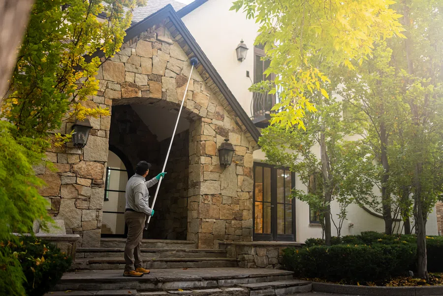 Person cleaning outdoor light fixture on stone porch of house surrounded by trees in daylight.
