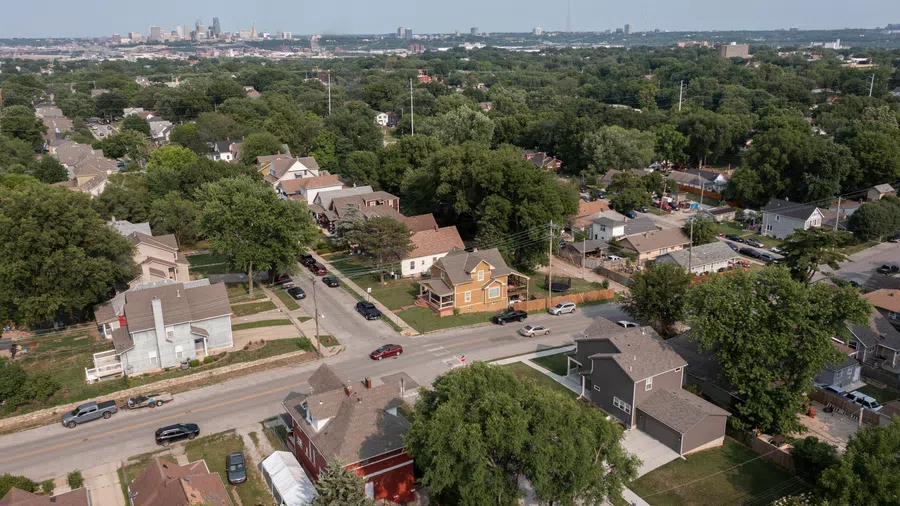 Aerial view of a suburban neighborhood with houses, streets, trees, and a city skyline in the distance on a clear day