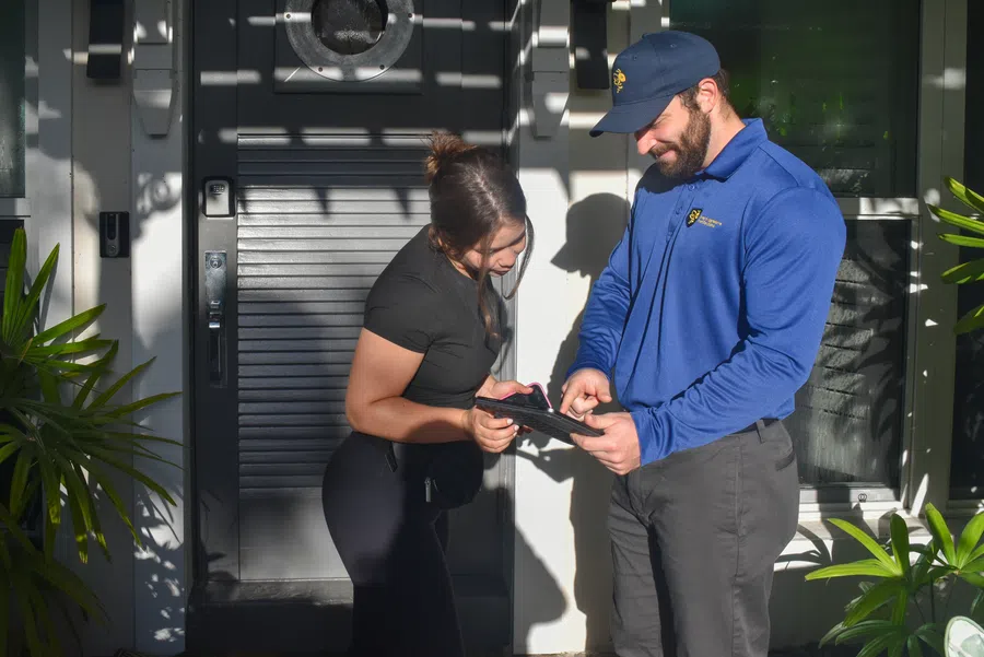 Delivery worker in blue uniform using a tablet while woman receives package at home entrance.