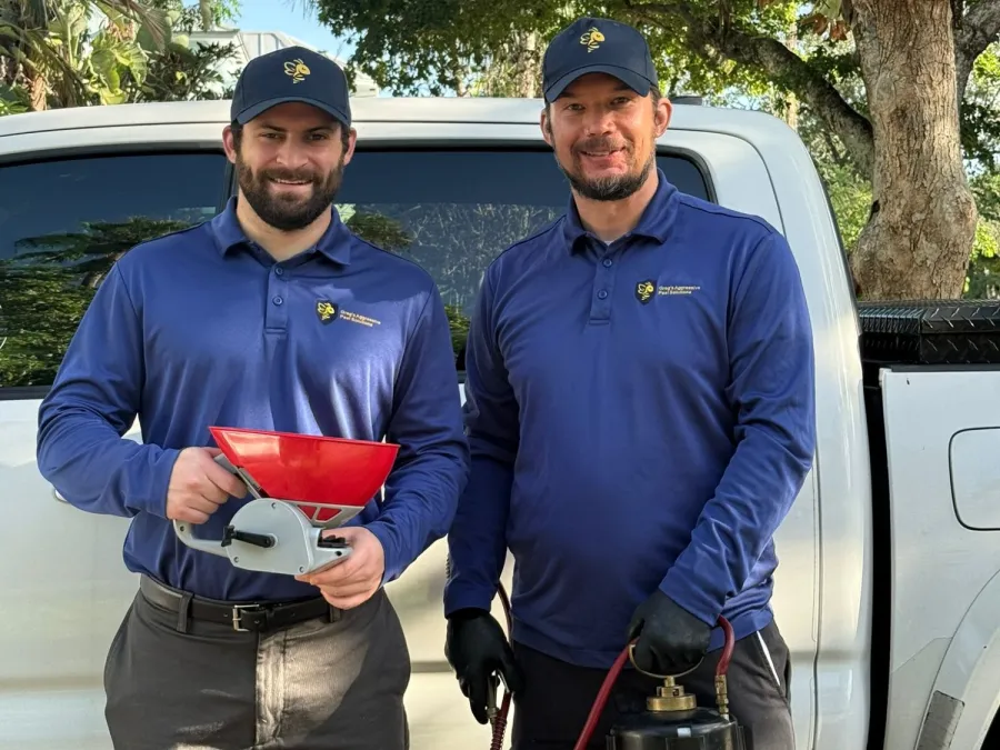 Two pest control workers in navy uniforms standing by a white truck holding pest control equipment outdoors.