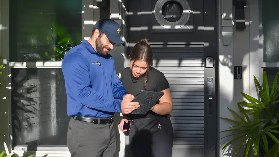 Delivery person in blue uniform shows a tablet to a woman outside a modern home entrance in daylight.