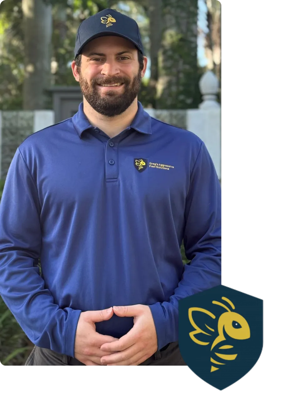 Man in blue Greg's Aggressive Pest Solutions uniform and cap smiles outdoors with green foliage background