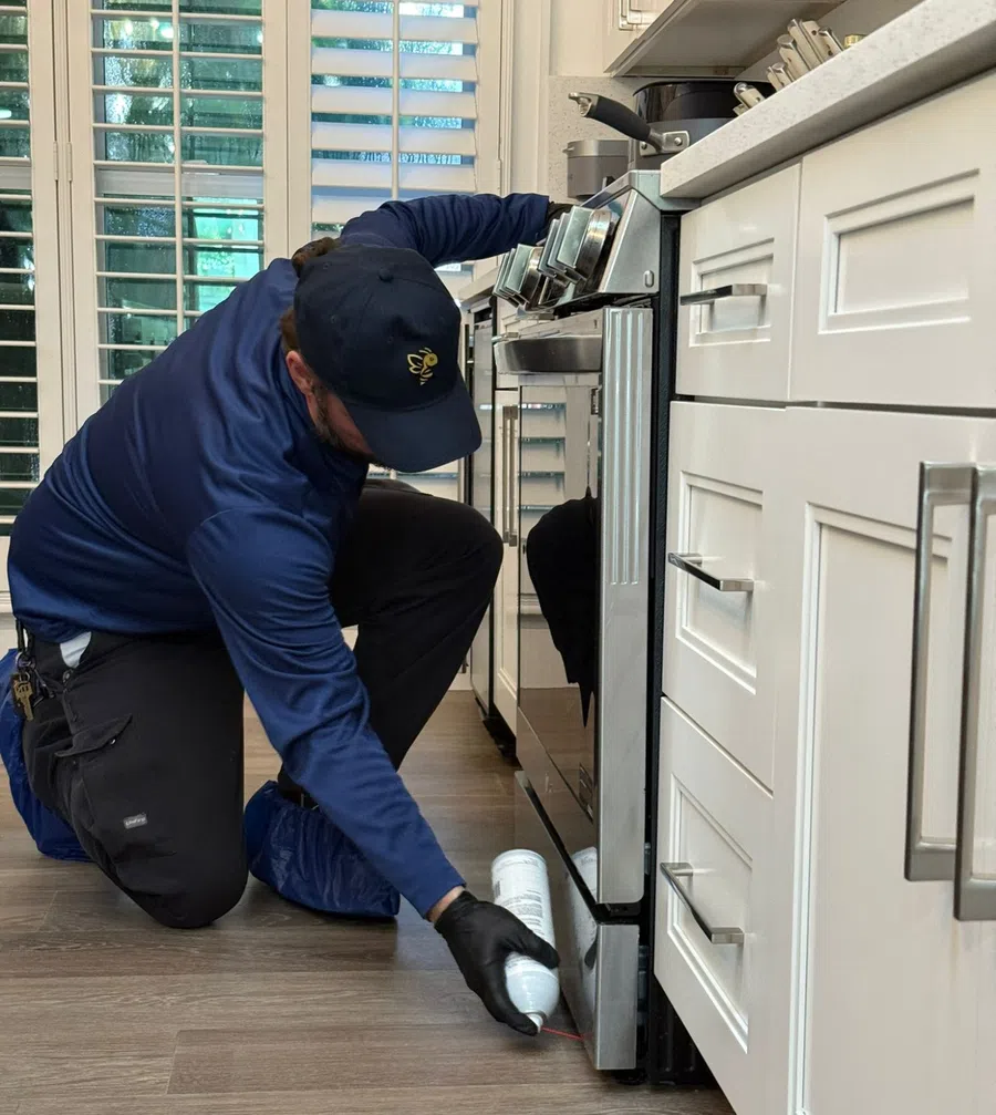 Technician in blue uniform applying pest control spray near kitchen stove on wood floor.