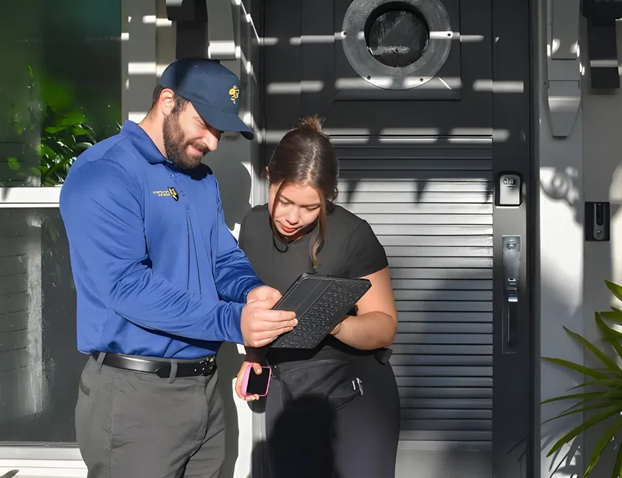 Delivery person in blue uniform shows a tablet to a woman outside a modern home entrance in daylight.