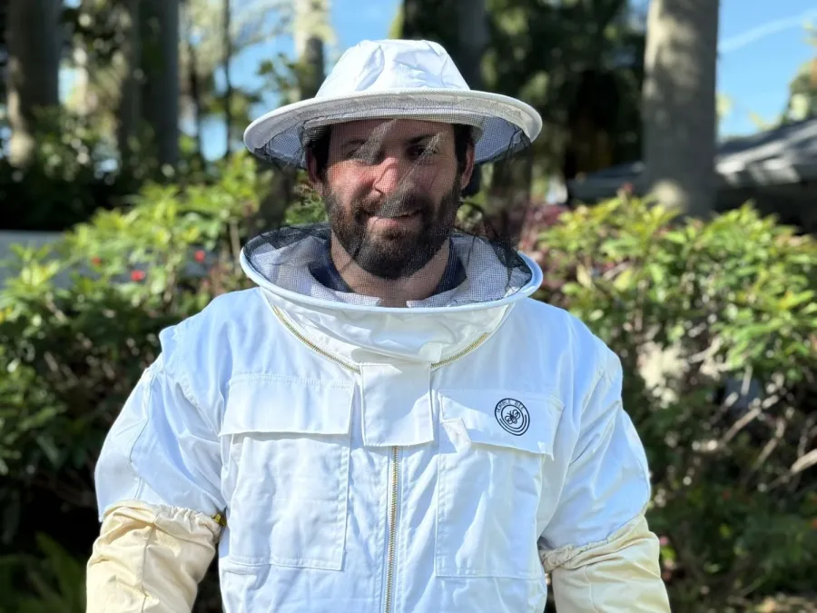 Man wearing a white beekeeping suit and yellow gloves standing outdoors with greenery in background