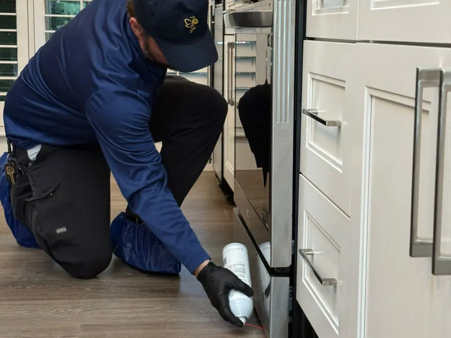 Technician in blue uniform applying pest control spray near kitchen stove on wood floor.