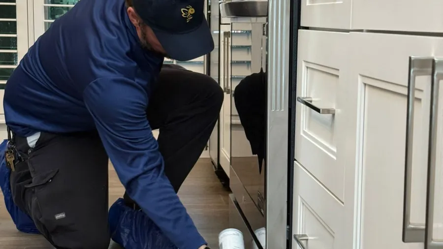 Technician in blue uniform kneeling on wood floor applying pest control spray near kitchen cabinets and oven.