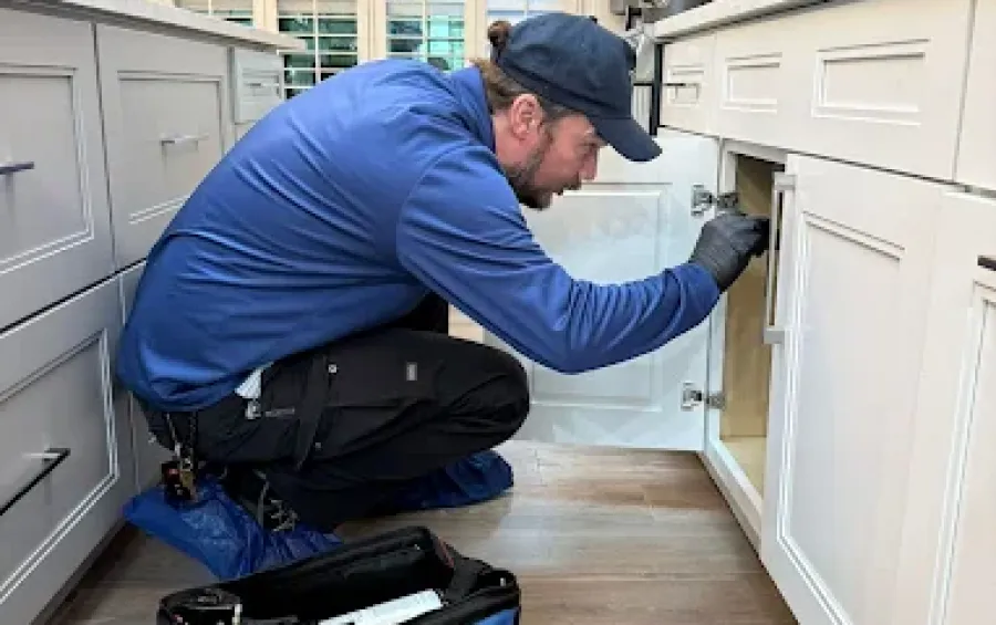 A plumber in blue uniform fixing a kitchen under-sink cabinet with a tool kit on the floor nearby.