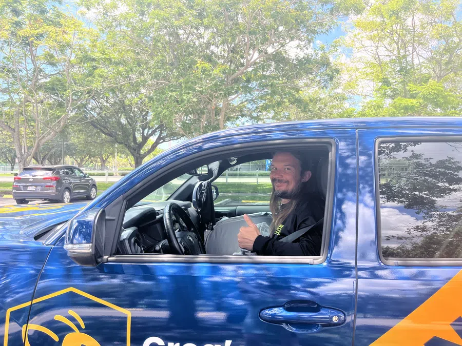 Man smiling and giving thumbs up inside a blue pickup truck with yellow logo parked by trees.