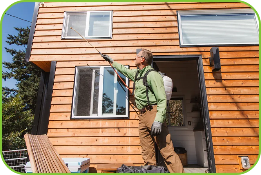 Man in protective gear spraying insecticide on a wooden house exterior on a sunny day.