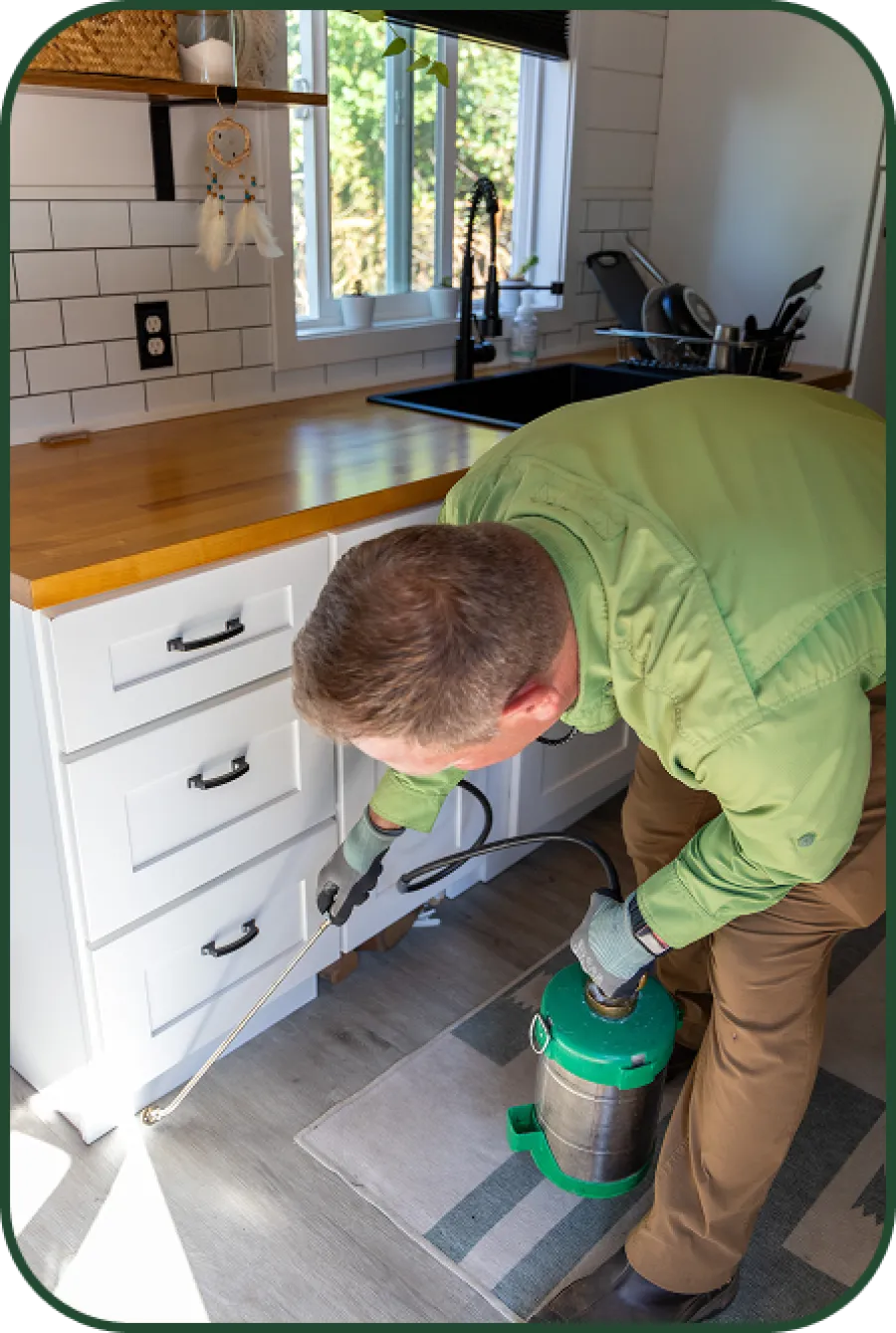 Exterminator in green shirt treating kitchen floor near white cabinets for pests with spray equipment.