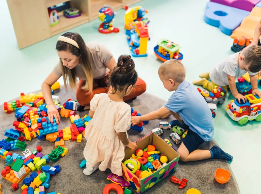 Children and female caregiver playing with colorful building blocks and toys on a classroom carpet.