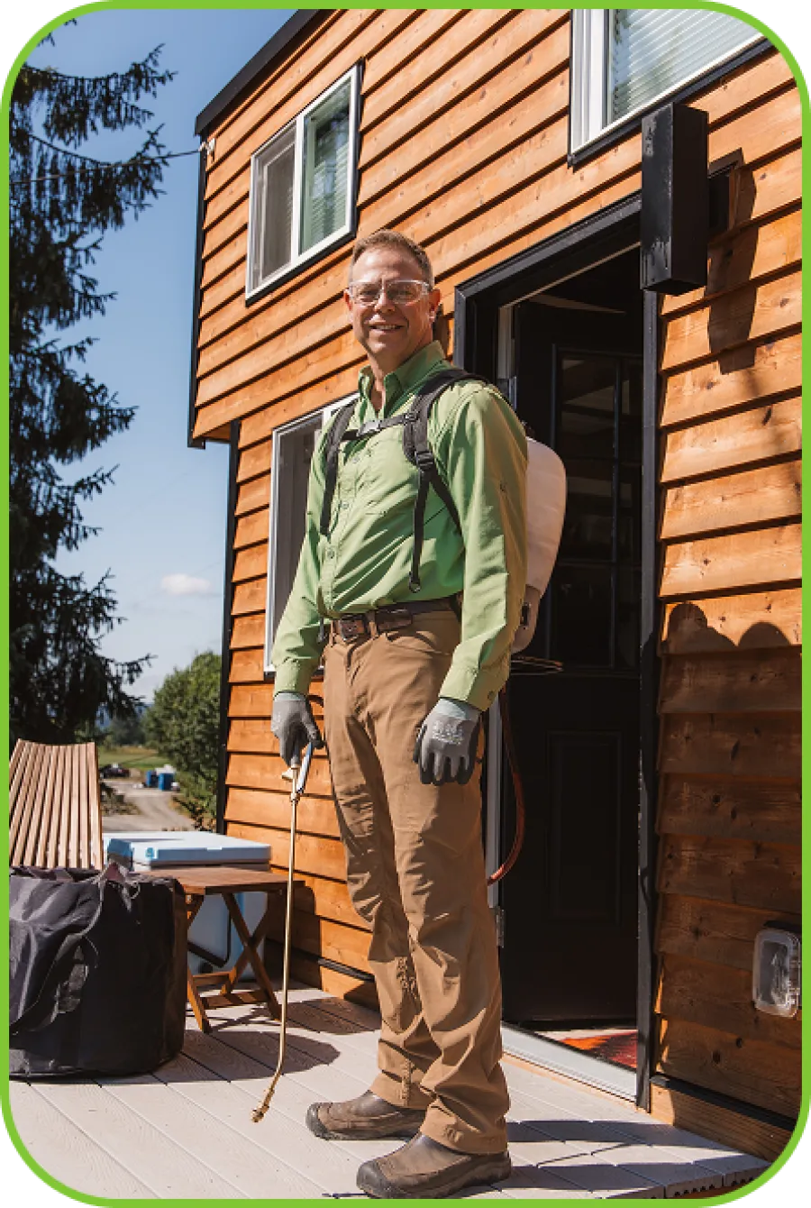Man in protective gear with backpack sprayer standing on wooden porch outside a house with natural wood siding.