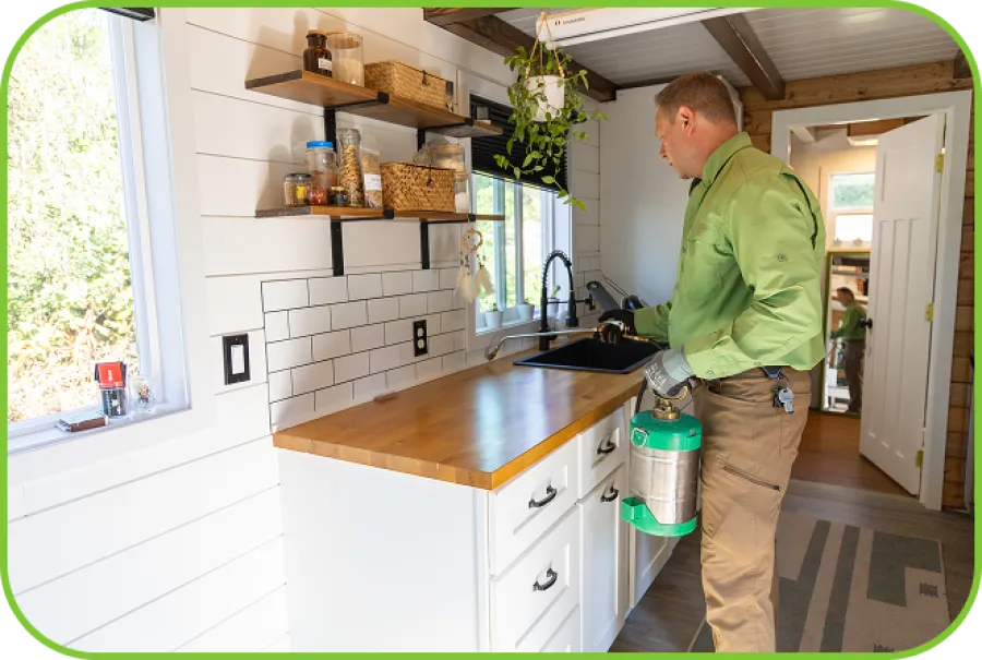 Pest control technician spraying insecticide under kitchen sink in a modern white kitchen.