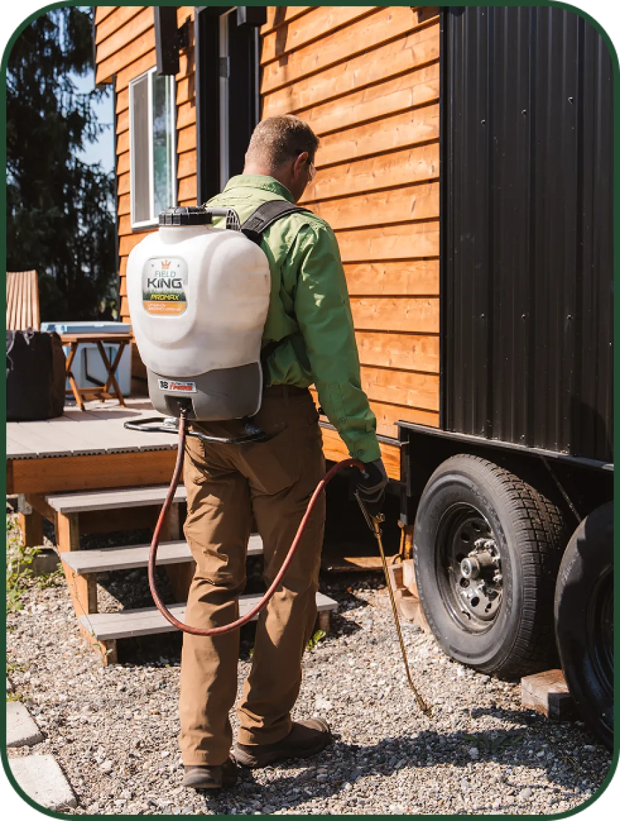 Man wearing a backpack sprayer applying pesticide or herbicide near a wooden house and trailer outdoors