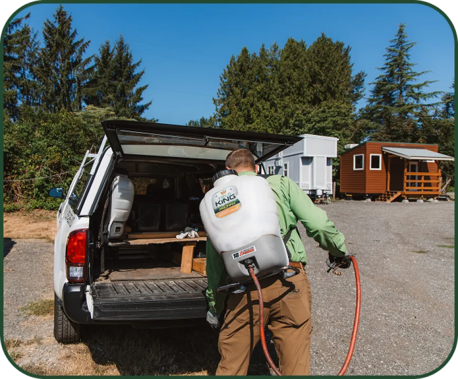 Man using backpack sprayer to apply treatment near wooden house and trailer on gravel surface.