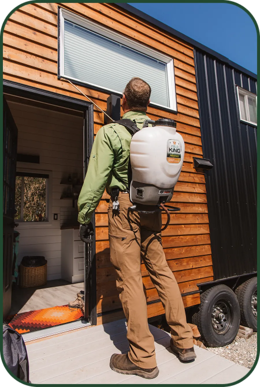 Man wearing backpack sprayer treating exterior of tiny house with wood siding and black metal paneling.