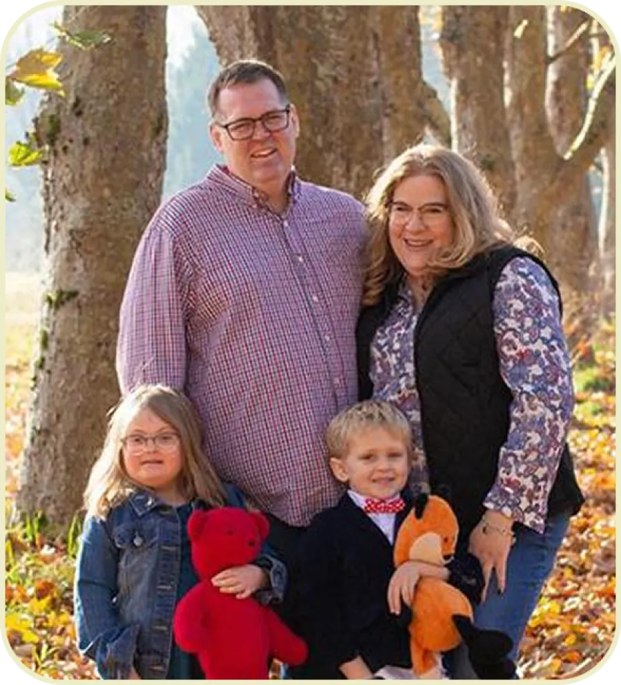 Smiling family of four standing outdoors in fall with two children holding plush toys among trees and autumn leaves